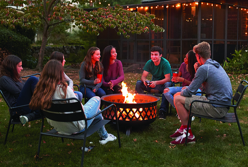 Teenagers sit talking around a fire pit in a garden at dusk